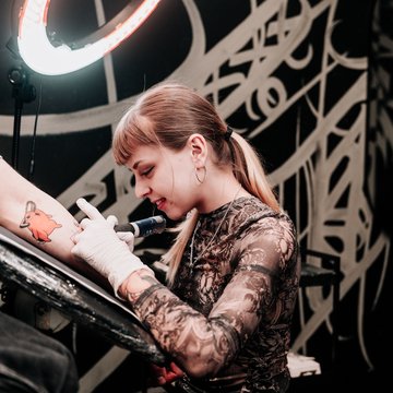Experienced tattoo artist at VEAN TATTOO in Tallinn tattooing a client’s arm under a ring light, wearing gloves in a clean, sterile studio focused on custom designs.
